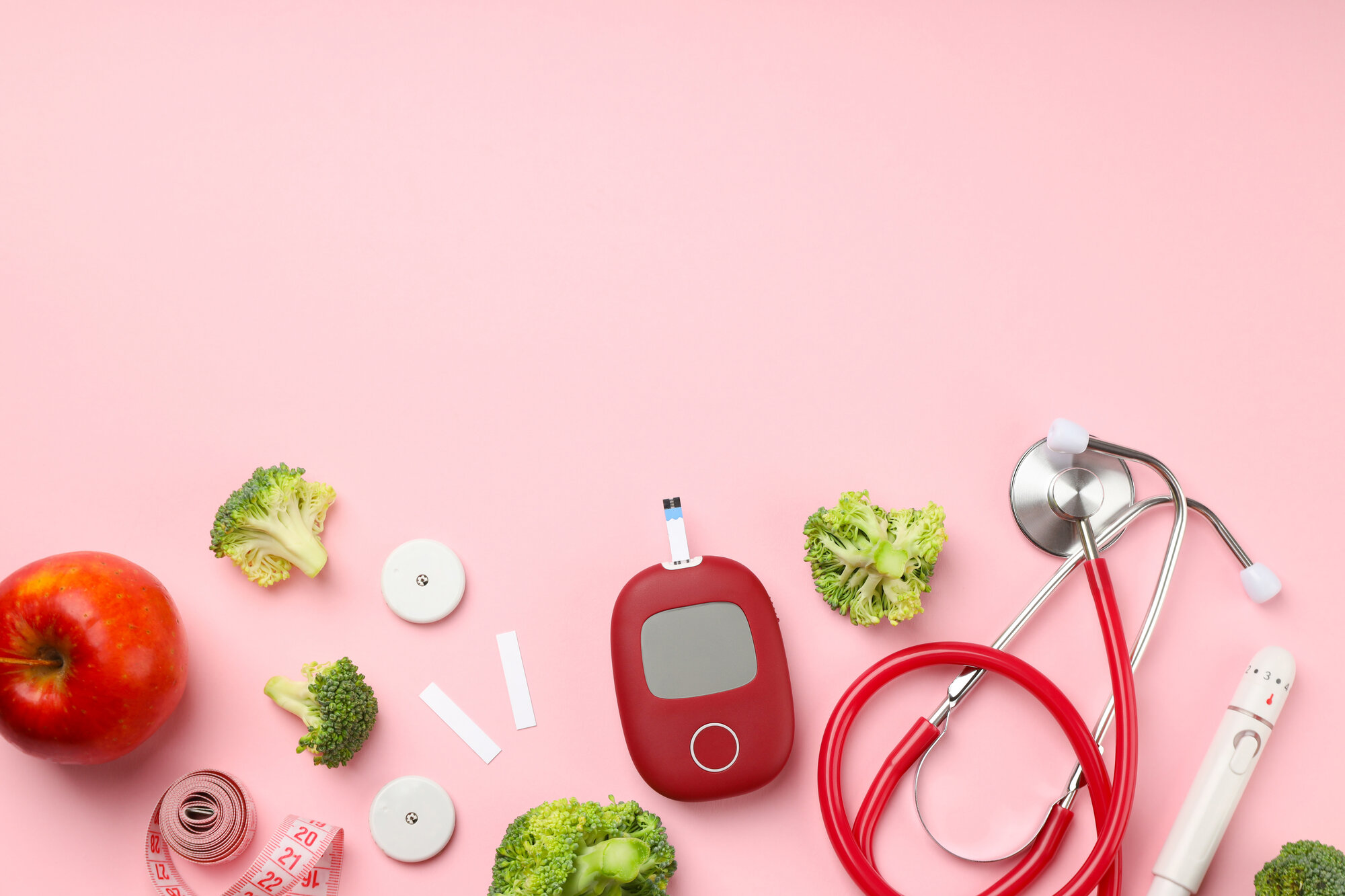 Blutzucker-spitzen.jpeg Gesunde Lebensmittel und medizinische Geräte auf rosa Hintergrund, darunter Apfel, Brokkoli, Blutzuckermessgerät und Stethoskop.