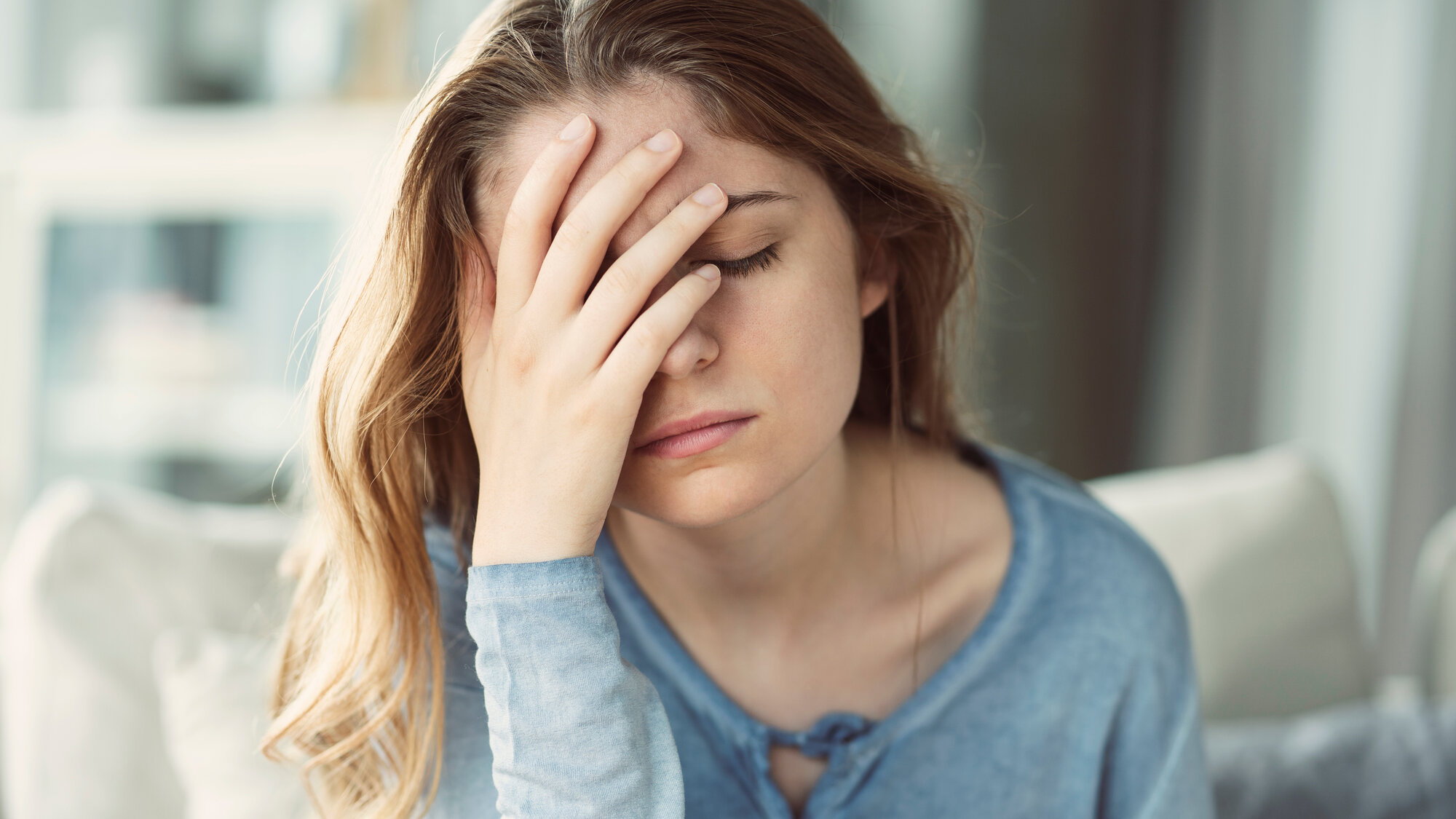 chronische-entzuendungen.jpeg Junge Frau mit langen Haaren und blauem Oberteil, die frustriert ihre Hand an die Stirn legt, im Innenraum sitzend.