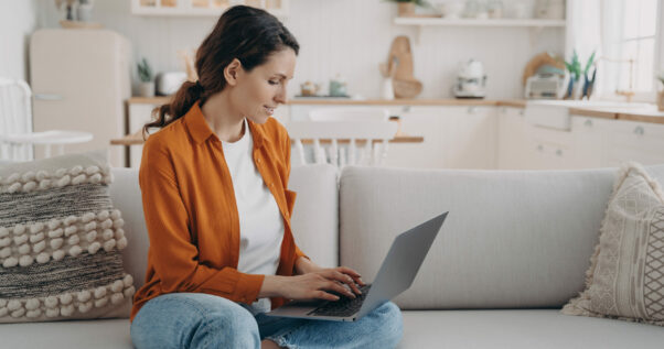 Woman is typing on pc and sitting on couch at home. European girl in orange shirt is chatting and browsing internet. Remote work or education. Businesswoman at cosy living room.